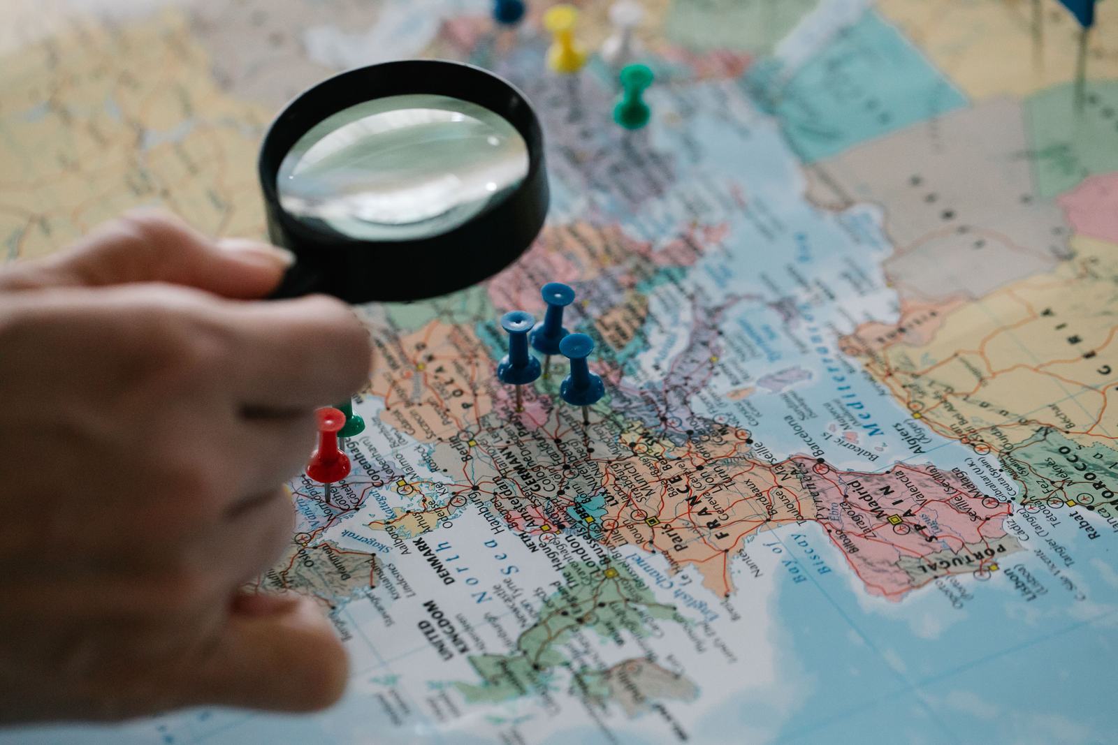 A hand holding a magnifying glass over a map with push pins, standing in for spotting a fake competitor listing crowding the Map Pack.