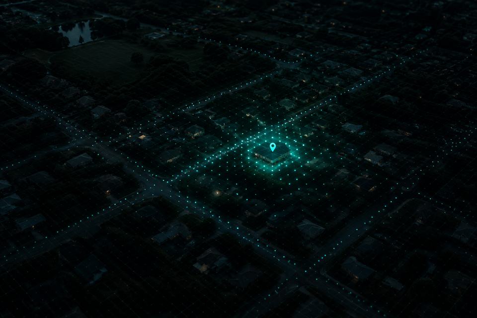 A night aerial view of a neighbourhood with a grid of glowing points, bright and dense around one central building and fading to nothing toward the edges.