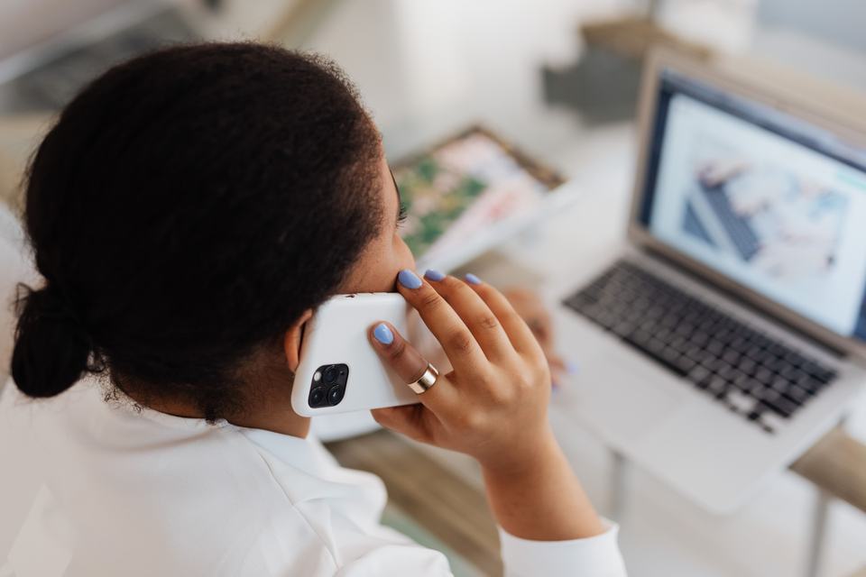 A woman holding a phone to her ear beside an open laptop, standing in for a customer who researches online and then phones